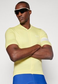 Kurzärmliges Fahrradtrikot in Hellgelb mit einem Reißverschluss an der Vorderseite, kombiniert mit blauen Shorts. Mit einem weißen Akzentband am Ärmel.