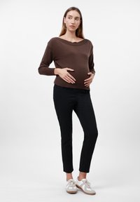 Pregnant woman in brown sweater and black pants standing with hands on belly, looking to the side against a plain white background.