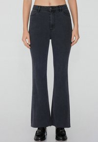 Hochgeschnittene schwarze, ausgestellte Jeans mit glatter Denim-Oberfläche, leichten Ausfransungen am Saum und standardmäßiger Fünf-Taschen-Optik. Schwarze Schuhe sichtbar.