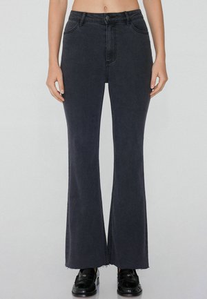 Hochgeschnittene schwarze, ausgestellte Jeans mit glatter Denim-Oberfläche, leichten Ausfransungen am Saum und standardmäßiger Fünf-Taschen-Optik. Schwarze Schuhe sichtbar.