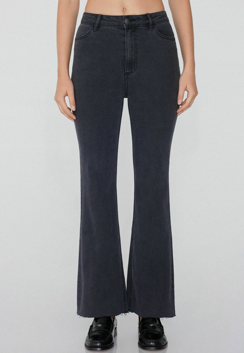 Hochgeschnittene schwarze, ausgestellte Jeans mit glatter Denim-Oberfläche, leichten Ausfransungen am Saum und standardmäßiger Fünf-Taschen-Optik. Schwarze Schuhe sichtbar.