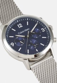 Reloj con correa de malla plateada y esfera de color azul marino, que presenta marcadores de hora blancos, tres subesferas y el logo de "Calvin Klein".