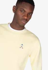 Sudadera amarilla clara con cuello redondo y un diseño de avión bordado en el pecho. Tejido suave, corte casual y mangas largas.