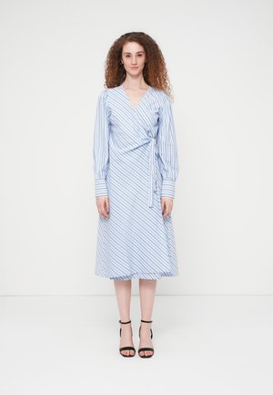 Femme aux cheveux roux bouclés portant une robe portefeuille rayée bleu clair et blanc, ainsi que des escarpins noirs à bride cheville, debout devant un fond blanc.