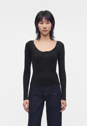 Ung kvinde med glat sort hår iført en tætsiddende sort langærmet top med blondekant og mørkeblå vidde jeans, stående mod hvid baggrund.