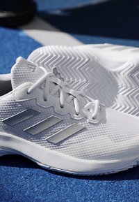 White sports shoes with textured mesh design and three dark stripes on the sides resting on a blue textured surface.