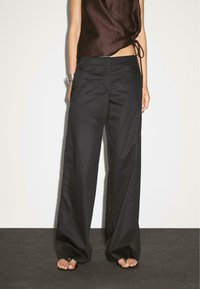 Le modèle porte un haut sans manches marron foncé et un pantalon noir ample, se tenant contre un arrière-plan clair, mettant en valeur des chaussures élégantes.