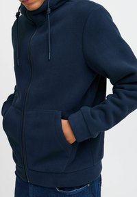 Marine fleece zipjack met dubbele voorzakken, verstelbare trekkoordkap en ribgebreide manchetten; zachte textuur en moderne pasvorm.