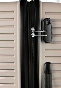 Maleta rígida beige con cremallera negra, doble tirador de cremallera plateado asegurado con un candado de combinación negro y un asa lateral negra.