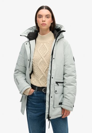 Light gray padded jacket with a black hood, multiple pockets, and a smooth texture, worn over a cream knit sweater and blue jeans.