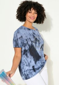 Blaues Batik-T-Shirt mit kurzen Ärmeln, das ein mit Strasssteinen besetztes Smiley-Gesicht in der Mitte zeigt, kombiniert mit weißen Hosen.