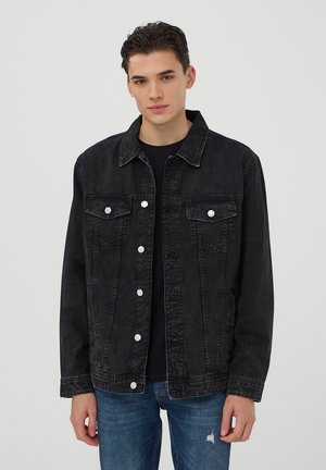 Giacca in denim nero con taglio classico, bottoni frontali, due tasche sul petto e tasche laterali. Tessuto leggermente sbiadito, abbinata a jeans blu.