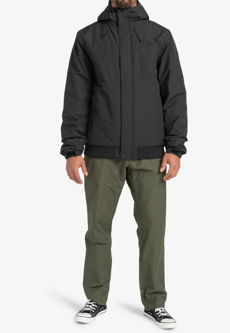 Sort isoleret jakke med hætte, elastiske manchetter og frontzipper. Sammen med olivengrønne bukser og sorte sneakers.