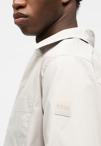 Κοντινό πλάνο ενός ατόμου που φοράει ένα ανοιχτό μπεζ σακάκι με ένα έμβλημα "BOSS" στο πάνω μέρος του βραχίονα, με έμφαση στον ώμο και το κάτω μέρος του προσώπου.