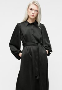 Blond naine, kes kannab pikk mustast satiinist kleiti, millel on punnis varrukad, nööbiga esiosa ja kinni seotud vöökoht, käed taskutes, neutraalne taust.