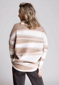 Beige- und weißgestreifter Pullover, weicher Stoff, lockere Passform, lange Ärmel, abgerundeter Saum, getragen mit dunkelgrauen Hosen, von hinten betrachtet.