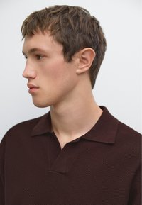 Polo shirt di colore marrone scuro con texture, dotato di colletto piatto e scollatura a metà. Modello in profilo, che mette in risalto il design della camicia.
