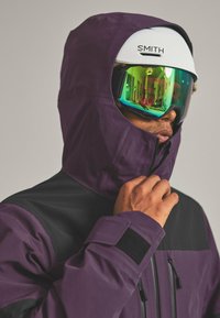 Ski jacket in purple and black, featuring a high collar, zippers, and adjustable hood. Includes reflective goggles with green lenses.