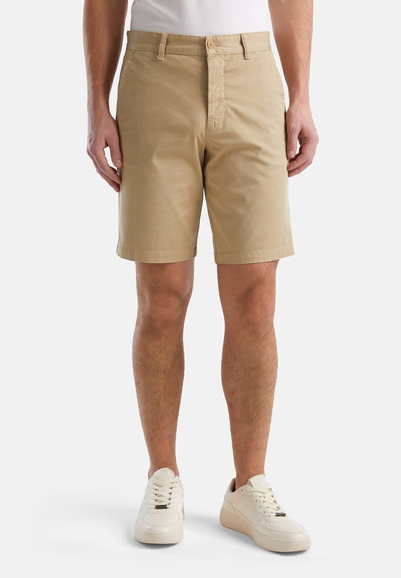 Shorts en coton beige avec une fermeture à bouton à l'avant, des passants de ceinture et des poches. Portés avec des baskets blanches. Texture lisse, coupe ajustée.