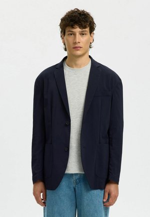 Mladý muž s krátkymi kučeravými vlasmi, oblečený v námornícky modrom saku cez svetlosivú tričko a modré džínsy, stojaci pred jednoduchým bielym pozadím.