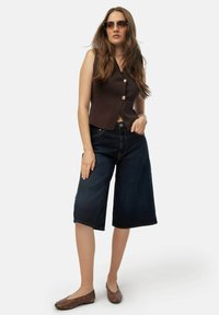 Bruine mouwloze vest met een diepe V-hals en gouden knopen, gecombineerd met donkere denim culottes en bruine studschoenen. Duidelijke zonnebril gedragen.