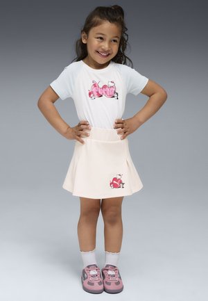 Lächelendes Mädchen mit Händen in den Hüften, das ein weißes Puma T-Shirt, einen beigen Rock mit Hello Kitty-Design, weiße Socken und pinke Puma-Sneaker trägt.
