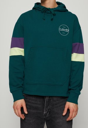 Mężczyzna w turkusowej bluzie z kapturem z fioletowymi i kremowymi paskami na rękawach oraz przedniej kieszeni, w połączeniu z czarnymi dżinsami.