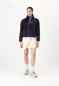 Svart fleecepullover med ett texturerat mönster och dragkedja i kvartslängd, ihopparad med en ljus grädds wrapkjol och grå sneakers.