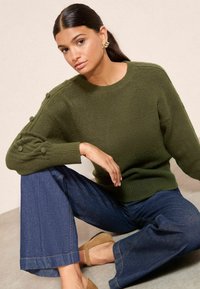 Pulover tricotat de culoare verde măslin, cu accente texturate pe mâneci, asociat cu pantaloni evazați din denim închis și saboți din suede bej.