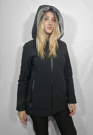 Giovane donna con lungo capelli biondi che indossa un giubbotto invernale nero con cappuccio, rifinito in pelliccia grigia, in piedi contro uno sfondo chiaro e semplice.