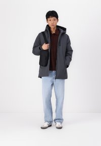 Mørk grå dunjakke med hætte, vist over en bordeaux top. Lyseblå, loose-fit jeans og en lille sort crossover taske. Hvide sneakers.