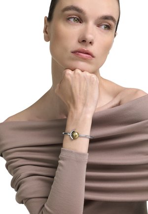 Tânără cu piele ușor măslinie și ochi verzi, purtând un top taupe cu umerii goi și o brățară argintie cu un charm sferic auriu.