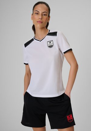 Kobieta o ciemnych włosach, ubrana w biały sportowy T-shirt z czarnym obszyciem i logo oraz czarne spodenki z czerwonym logo, stoi z rękami w kieszeniach.