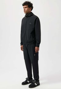 Costum de trening ușor, de culoare neagră, cu jachetă cu glugă, manșete elastice și pantaloni asortați cu un buzunar lateral. Asortat cu adidași negri.