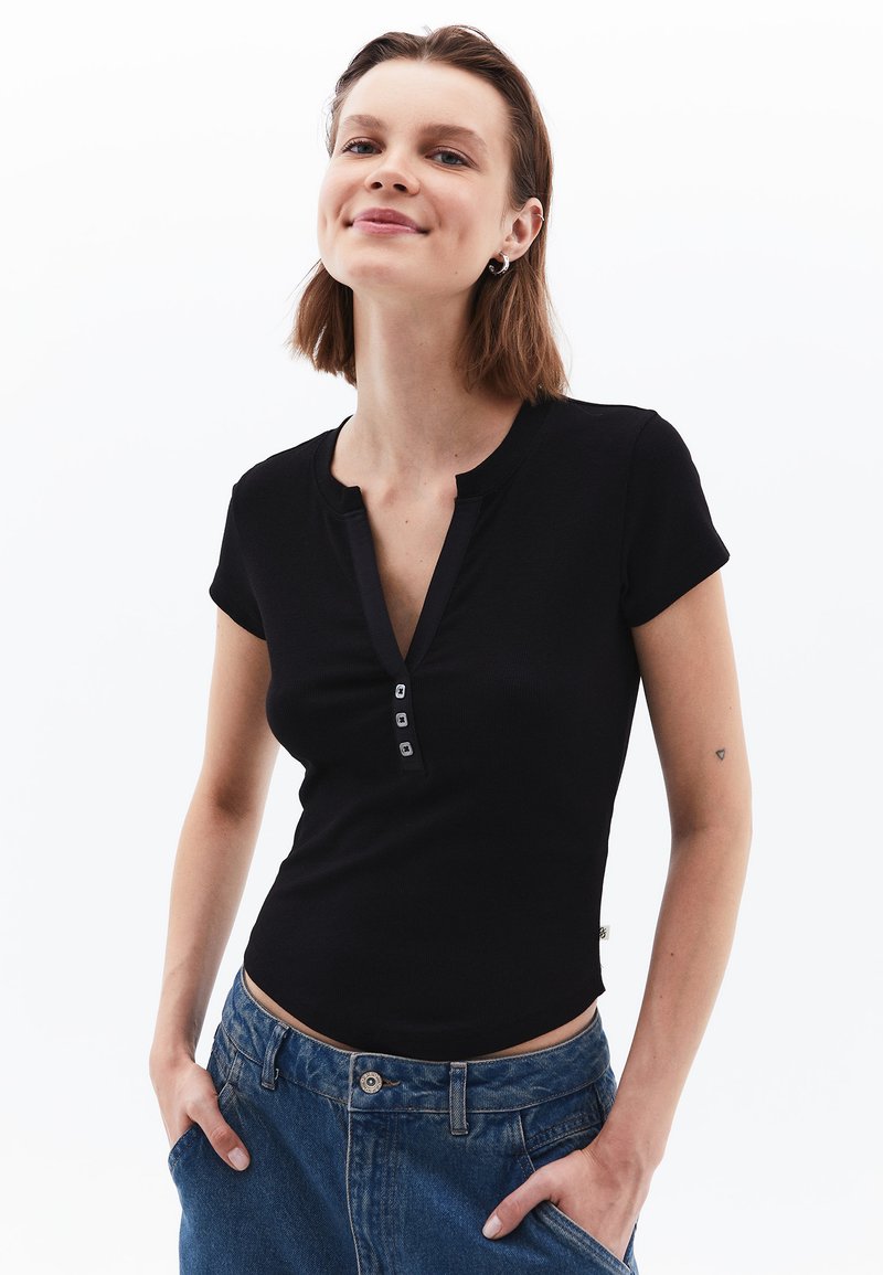 Sort kortærmet top med ribbet tekstur, featuring en V-udskæring og fire-knap lukning. Båret sammen med høj tale denimbukser.