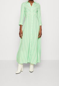Robe longue boutonnée de couleur vert clair avec un motif de fougère blanche. Elle possède un col et des manches trois-quarts, portée avec des bottines blanches à talons blocs.