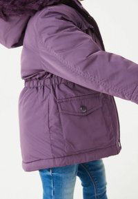 Veste matelassée violette avec taille cintrée, poches avant et capuche amovible en fourrure. Texture lisse ; design ajusté, isolation chaude.