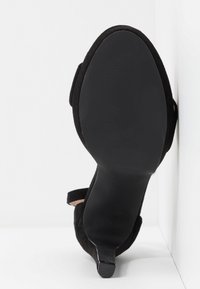 Vue de dessous d'une chaussure à talon noir avec un talon pointu et une texture en daim sur une surface blanche.