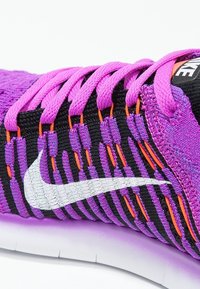 Zapatilla de Nike con parte superior de tejido morado, rayas negras, acentos naranjas y logotipo blanco. Presenta cordones rosas y suela blanca. Superficie texturizada.