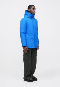 Blue padded jacket with a high collar, two chest pockets, smooth finish, paired with loose black cargo pants and embellished slip-on shoes.