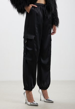 Pantaloni cargo in satin nero con vita elastica, tasche laterali e polsini arricciati, abbinati a scarpe con tacco alto in argento.