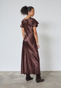 Kvinna med lockigt hår i en hästsvans bär en glansig brun maxiklänning med korta ärmar och mörkbruna stövlar, stående på betonggolv.