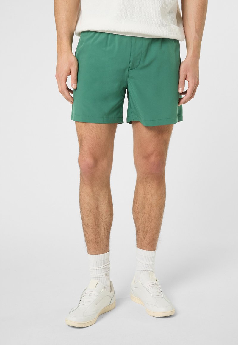 Homme portant un short vert, des chaussettes blanches et des baskets blanches, debout devant un fond clair uni.