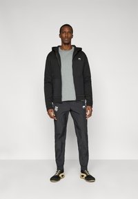 Μαύρο φούτερ με φερμουάρ μπροστά, γκρι πουκάμισο από μέσα και μαύρο αθλητικό παντελόνι με λευκές λεπτομέρειες. Συνδυασμένο με μαύρα και μπεζ παπούτσια.