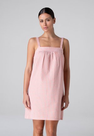 Donna con capelli scuri che indossa un vestito senza maniche rosa e bianco a quadretti, in piedi davanti a uno sfondo grigio chiaro uniforme.