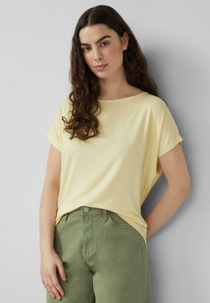 Młoda kobieta z długimi kręconymi włosami, ubrana w luźny, bladożółty top i wysokie oliwkowozielone spodnie, stoi na tle szarej ściany.