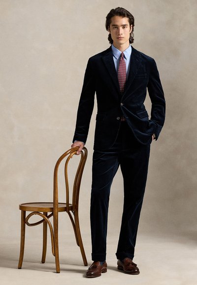 Παλτό από βελούδο σε σκούρο μπλε χρώμα με εφαρμοστή γραμμή, σε συνδυασμό με γαλάζιο πουκάμισο και γραβάτα μπορντό με σχέδια. Καφέ παπούτσια. Ξύλινη καρέκλα κοντά.
