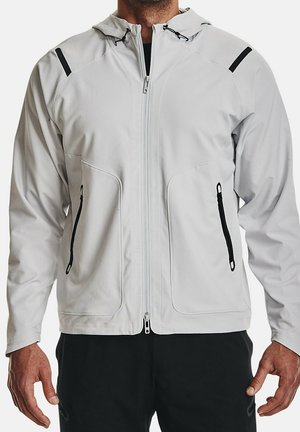 Mann trägt eine hellgraue Kapuzenjacke mit Frontreißverschluss und schwarzen Reißverschlusstaschen, kombiniert mit einer schwarzen Hose, steht vor weißem Hintergrund.