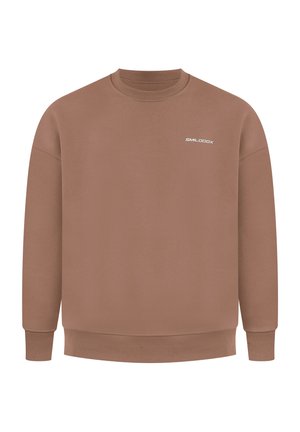 Mężczyzna w brązowej bluzie z białym nadrukiem na plecach, jasnoszarych spodniach dresowych i białych butach sportowych, stoi i patrzy w dół.