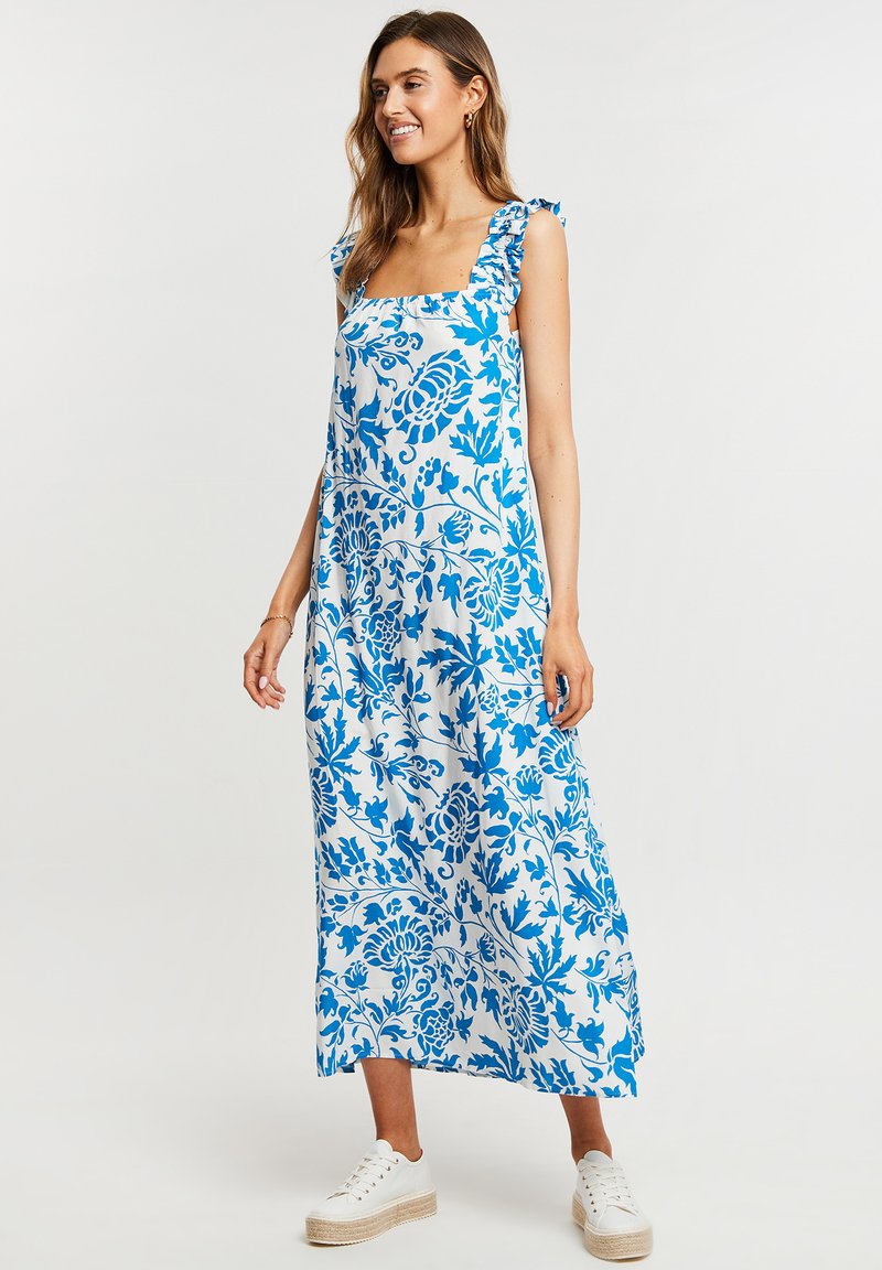 Maxi vestido de estampado floral en azul y blanco, con tirantes de hombro con volantes, escote cuadrado y aberturas laterales, combinado con zapatillas blancas.
